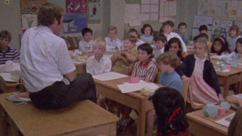 A classroom of children sit around a man on a teacher's desk.