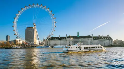 The London Eye and the former County Hall pictured from the other side of the River Thames. There is a boat going past on the river and it is a sunny day with blue skies. 