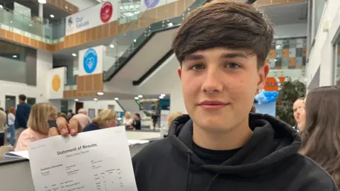 Ethan is holding up a piece of paper which reads: "Statement of Results". He has short, brown hair, an earring and is wearing a black hoodie. He is smiling slightly at the camera.