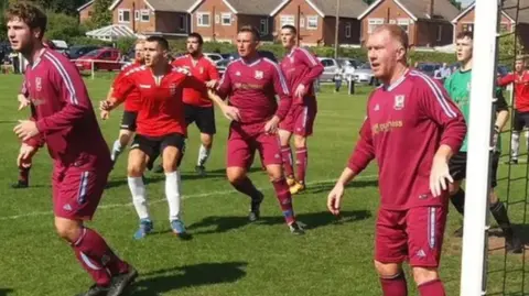 Peter Baldwin A group of players brace to intercept the ball on a football pitch mid-play. The players are dressed in burgundy kit. The man stood by the goal post is Paul Scholes. There are a number of residential houses to the right of the pitch. 