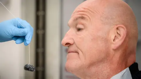 Cancer Research UK A man with a bald head looks at a small black sponge on a string being held by a hand wearing a blue glove