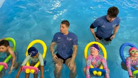 A group of children kitted out in inflatable swimming training kit holding the bar at the edge of a pool with an instructor next to them