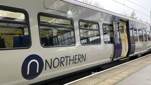 An image of the side of a Northern train as it pulls up at a station. The company's logo can be seen in blue lettering and the train's doors are open. No passengers can be seen seated in the windows.