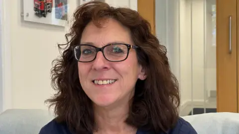 A woman with curly brown shoulder length hair and black glasses, wearing a navy top