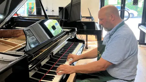 Adam Cox, Yorkshire Piano Piano Employer playing Piano's Black Piano in his space room. He wears a blue shirt with a green Apron. In the background, various pianos can be seen.