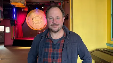 Rufus has dark hair and a beard and wears a checked red and blue shirt and a dark blue jack. He is standing in The Cornish Bank with the stage behind him. 