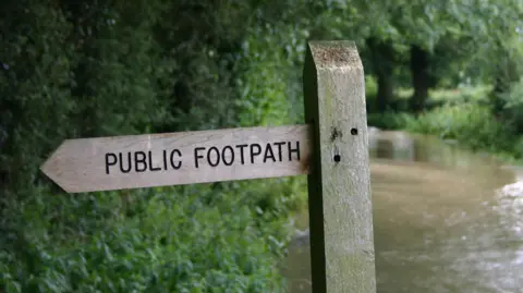 A wooden signpost, saying public footpath, with a stream and vegetation in the background.