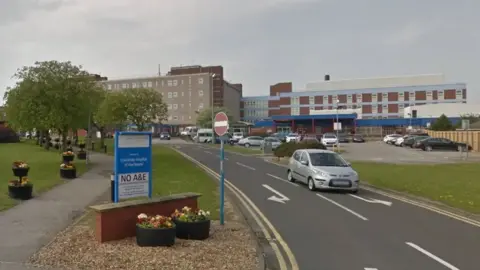 Entrance to University Hospital of Hartlepool. There is a patch of green grass on the verge of the road with an 80s-design hospital building in the background. 