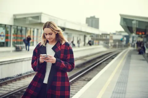 Getty Images A young woman with shoulder length blonde hair wearing a red and black checked coat walking along a railway platform looking at her phone