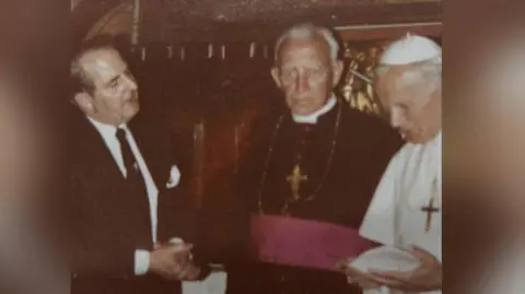 Family Photo Eddie Rabaiotti shows Pope John Paull II the menu for the dinner. The Pope is on the right, he is wearing white robes and a gold cross around his neck. Next to him is a bishop wearing a black robe and a white collar, he is wearing a gold cross. Eddie stands on the left with his hands clasped, he is wearing a black suit and a white shirt. There is a white flower in his label. 