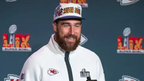 Travis Kelce smiles when answering a question into a mic. 