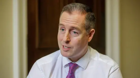 PA Media A man with short dark grey hair is talking to someone off camera. He is wearing a white shirt and purple patterned tie. The background is out of focus, but he sits in front of a large brown wooden door.