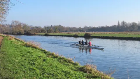 Four people in a boat heading upstream on a bright, cloudless day. The photograph has been taken from the Chesterton side of the River Cam, on a grassy embankment. 