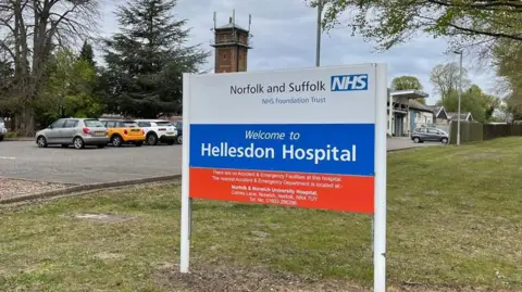 Jamie Niblock/BBC The picture is a close up of the sign for Hellesdon Hospital. It says Norfolk of and Suffolk NHS Foundation Trust is white red and blue. It also says "Welcome to Hellesdon Hospital". There is a car park behind the sign and some of the cars are visible. The sign it is positioned on grass.