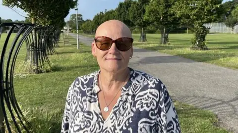 Phil Harrison / BBC A woman wearing a blue and white patterned top. She is stood in a park, in front of a tarmac path, surrounded by trees.