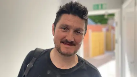 Simon Thake A man with dark hair and a beard smiles but without any teeth.