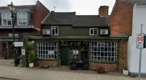 Google The bar is painted olive green and has large square windows either side of the entrance which has two seats and a table outside as well as some greenery. There are two levels to it.