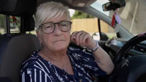 A woman with short grey hair, black round glasses and a blue-with-white-stripes patterned top sits in the driver's seat of a car. Her arm rests on the steering wheel, and she's wearing a bracelet. She looks concerned. The car's upholstery is black. There are purple and red air fresheners hanging from her rear view mirror.