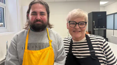 A young man and an older women, both wearing aprons, smile as they stand next to each other in a kitchen. Then man is wearing a grey jumper and a bright yellow apron. He a long brown hair and a full beard. On the right, the woman is wearing a striped white and black top, a black apron and black-rimmed glasses.