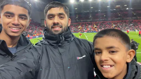 Ibrahim Syed Ibrahim Syed smiles for a selfie, flanked by his two sons in the stands at Anfield. Players warm up on the pitch behind.