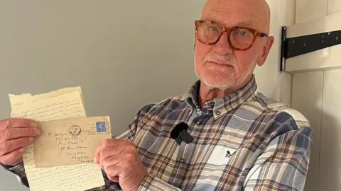A bald man with a white stubble and wearing red-rimmed glasses, holds a piece of paper with writing on it and an envelope up for the camera. The man stands next to a wooden door, painted white, and wears a white and brown tartan shirt.