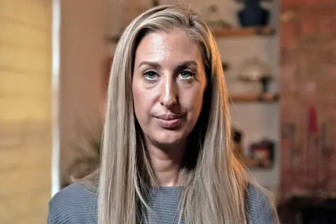 MARTIN MCQUADE / BBC Portrait photo of Fiona Winser-Ramm. She has long, straight blonde hair with a side parting. She has blue eyes and is wearing a grey ribbed jumper. She is pictured in a living room, with shelves and a blind visible behind her. 