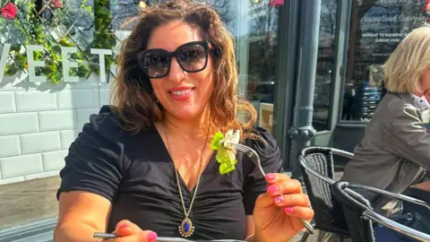 Vicki Broadbent Vicki sitting at an outdoor table eating a bowl of salad. She is holding a fork with a bite of food and has a cup of coffee, cutlery, and a menu on the wooden table. The restaurant’s exterior has large windows and decorative flowers are visible behind them.