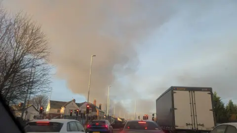 Plumes of billowing smoke in the sky, seen from traffic on a dual carriageway road.