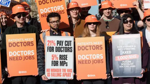 A group of resident doctors stand with orange signs saying 'doctors need jobs' and 'fund the NHS' outside parliament in London on Tuesday