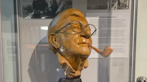 Chris Butterfield The plaster original of the sculpture of Alfred Wainwright made by Clive Barnard that the bronzes were cast from. The ochre-coloured sculpture featured the face of Wainwright with a pipe in his mouth. He also has a real pair of glasses over his face. The sculpture is inside a display cabinet.