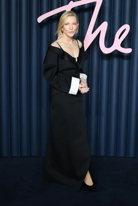 Getty A full-length view of Cate Blanchett in black dress with white sleeves, hand clasped together, standing in front of a blue curtain background