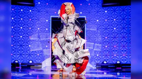 The image shows a drag queen in a dramatic dress made out of newspaper with red accents and tall heels. Her hair is up in a bin and red string can be seen in it. The backdrop is purple with white strobe lights.