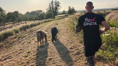 Handout Giles running on a country path through fields with trees and yellowed grass. He is joined by two small ponies on his left. He is wearing a branded cancer charity t-shirt and has his back to the camera.