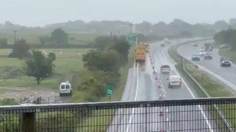 Cars drive down the A30 in Devon. One lane is closed off towards the exit slip road at Sourton Cross, near Okehampton. It is wet.