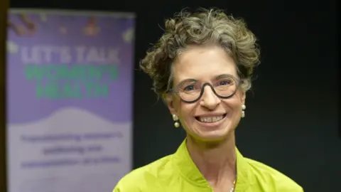 Dr Liz O'Riordan wearing a lime green outfit. She is looking directly at the camera and smiling. Behind her is a purple information board with lettering that says, LET'S TALK WOMEN'S HEALTH.