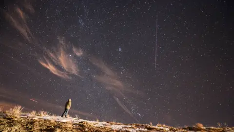 Getty Images Seseorang berdiri di punggung bukit yang tertutup salju pada malam hari sambil memandang ke langit, tempat garis meteor Geminid vertikal terang muncul di antara banyak bintang.