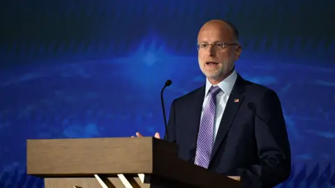 FCC chairman Brendan Carr speaking at a wooden podium wearing a navy suit, white shirt and red and blue checked tie.