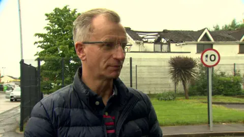 Brian Kingston is looking to the side. He wears glasses and has a blue jacket on with a red and blue stripped top. A building behind him as been damaged in a fire that's now out. 