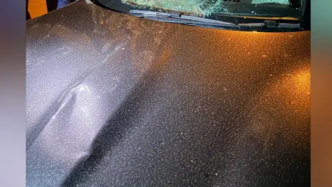 LDRS A badly smashed up windscreen on a black sports car. The bonnet has several large dents.