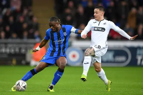 Getty Images Two football players clash over the ball - one dressed in blue and black stripes, the other in a white kit with hints of black. 