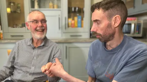 David Sincock (left) sitting next to his dad, Ian, in a kitchen. Ian is smiling at him as they bump fists. Ian has short grey hair and a beard and is wearing glasses along with a grey checked shirt. David has short dark hair and a beard. He is wearing a blue T-shirt. Behind them are cupboards filled with items of food, and a microwave can be seen on a worktop over David's shoulder.