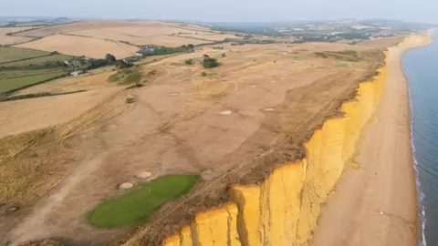Finnbarr Webster/Getty Images An aerial view of a clifftop and a golf course and fields to the left. The grass is brown due to drought and the sky is grey in colour. 