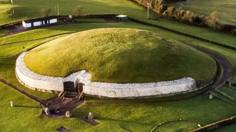 Ken Williams Newgrange - a mound passage tomb. It has a white stone frontage capped with a grass-covered dome. Grass surrounds it.
