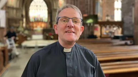 A man with grey hair and glasses, wearing a black shirt with a white dog collar. He is smiling at the camera. There are rows of pews behind him