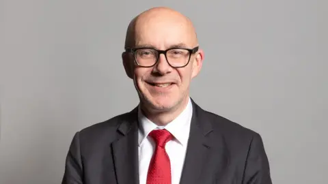 UK Parliament A man stands on a grey background. He is wearing a grey suit jacket, white shirt and red tie. He is bald and wearing glasses.