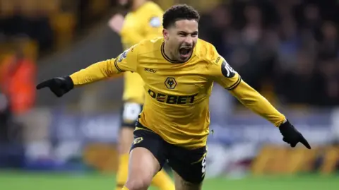 Getty Images Gomes is celebrating on the pitch. He has both arms outstretched and index fingers pointed and is shouting as he runs, possibly after scoring a goal.