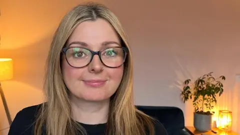 Head and shoulders of a bespectacled woman with long blonde hair and a nose ring woman at home, with lamps lit and a plant on a stand