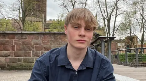 BBC/Marian McNamee Ben has blond hair and is wearing a dark blue shirt. He is sitting on some steps in front of a low sandstone wall, which itself is in front of a church.