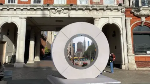 A computer generated image of what the portal could look like. It is a grey circle with a screen in the middle, it is on the Cornhill in Ipswich with buildings in the background. 