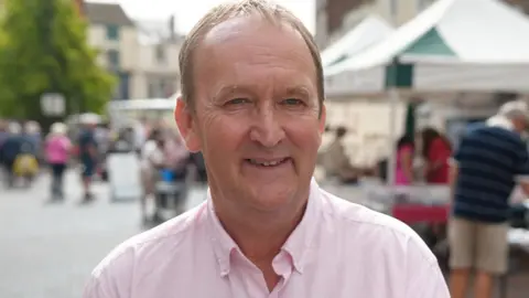 A man with short fair hair and wearing a pink Oxford shirt smiles at the camera as he stands in front of market stalls in a square.